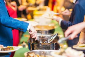 Guests are serving themselves food from a buffet table, using serving utensils and holding plates at what appears to be a wedding or formal event. The elegant setting hints at careful details, perhaps even taking place at the National Aquarium.