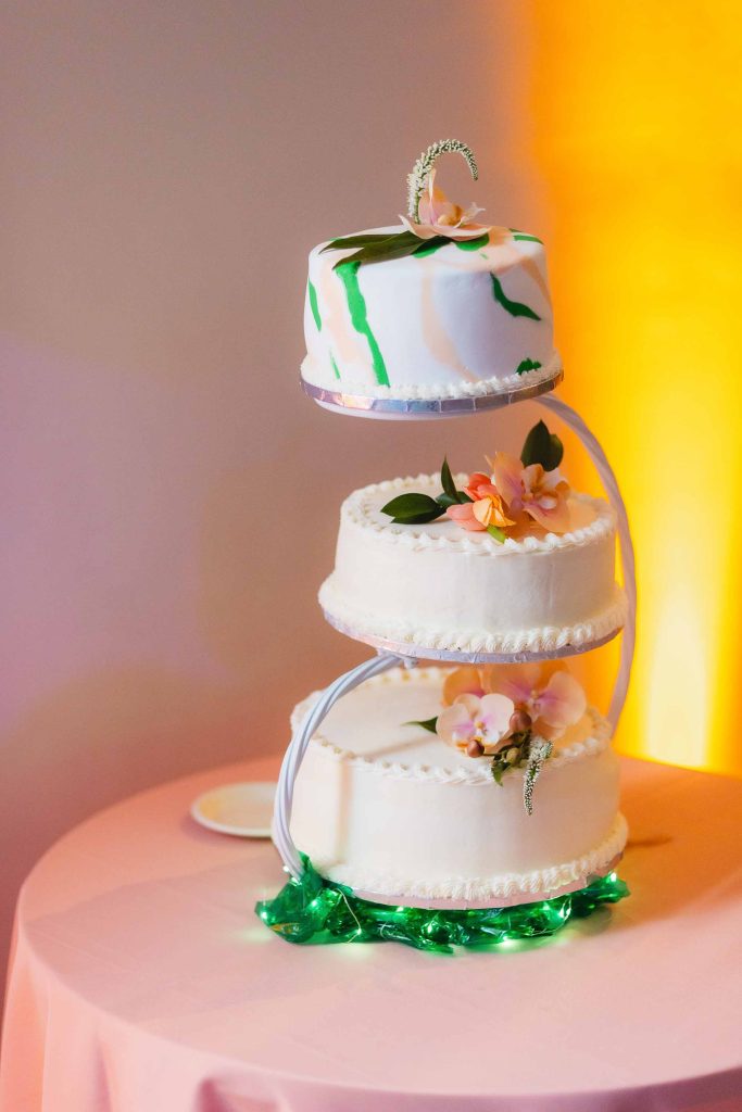 Three-tiered white wedding cake with floral details on a spiral metal stand, displayed on a round table with a pink tablecloth at the National Aquarium against a softly lit background.