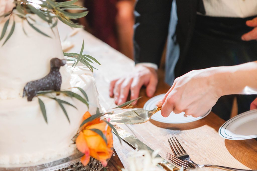 A person is cutting into a two-tiered white wedding cake adorned with green leaves and an orange flower during a wedding reception at The Barns at Hamilton Station. A small black dog figurine sits atop the cake while another person holds a plate.