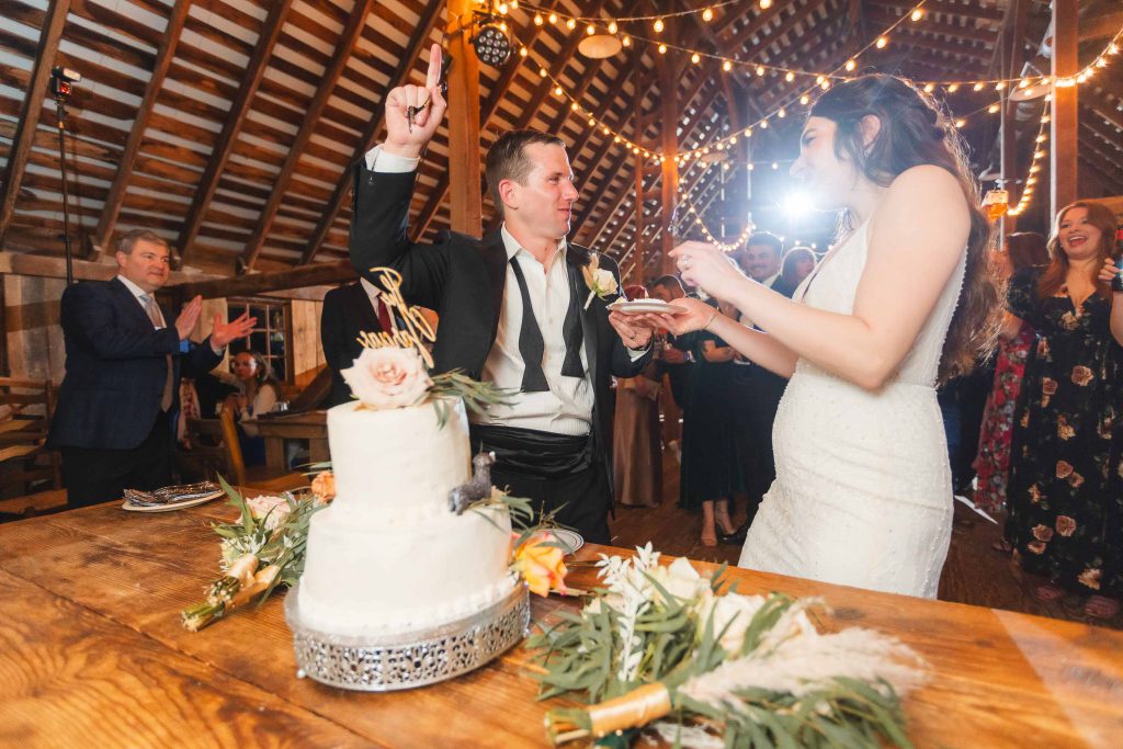 A newlywed couple is cutting a wedding cake at The Barns at Hamilton Station, a rustic, barn-style venue with string lights above. During the reception, the groom points upward while holding a piece of cake, and guests clap and smile in the background.