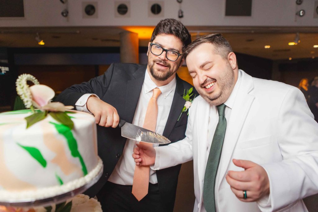 Two men stand together smiling at a reception as one, dressed in a black suit, cuts a decorated cake; the other man in a white suit looks on, laughing. The celebration unfolds at the National Aquarium in Baltimore.