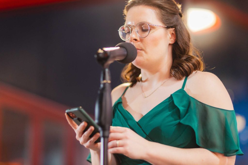 A woman in a green dress speaks into a microphone while reading from her phone during a wedding reception.