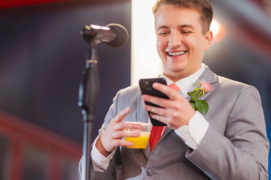 A man in a gray suit with a boutonniere holds a drink and a phone, smiling while standing in front of a microphone at a wedding reception.