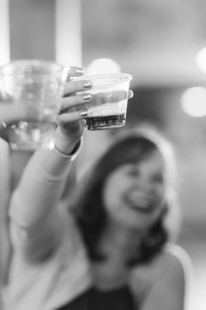 A person holds up a plastic cup in a toast at a wedding reception, with another cup nearby; the background is out of focus and the image is in black and white.