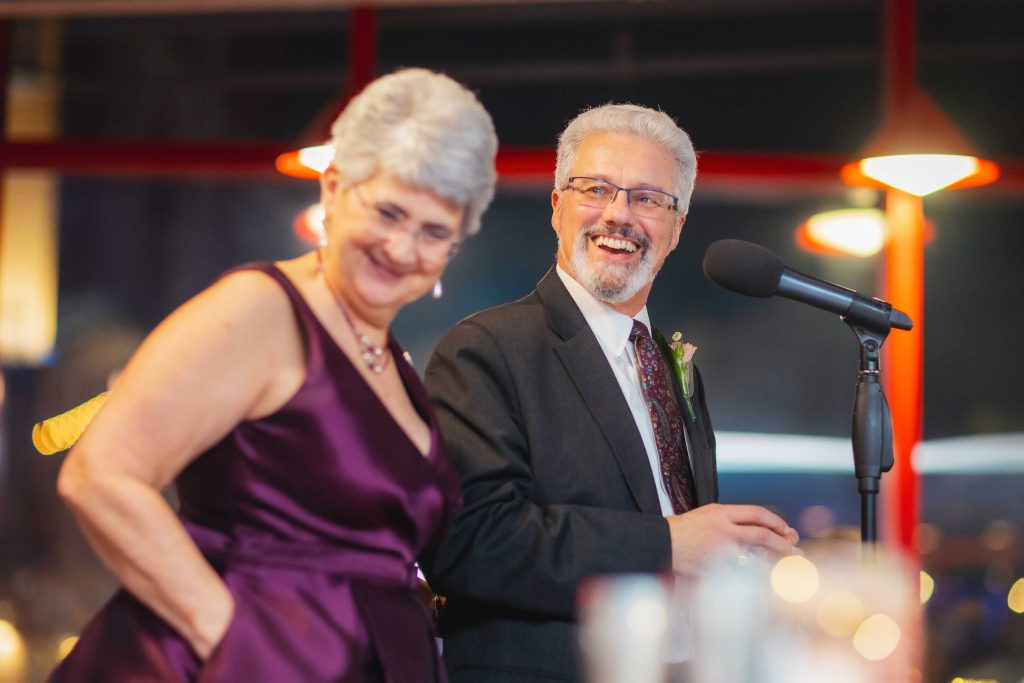 An older woman in a purple dress and a man in a suit stand near a microphone, smiling, at a wedding reception in Baltimore with warm lighting.