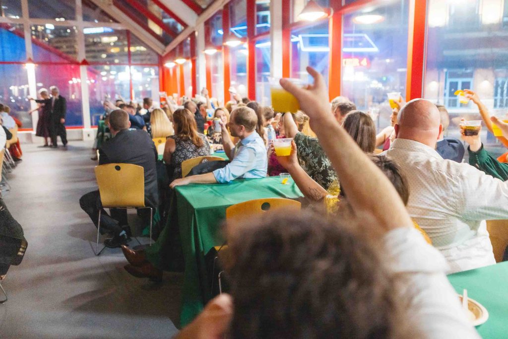 A group of people seated at tables indoors raise drinks in a toast while watching two speakers at the front of the room during a wedding reception at the National Aquarium.