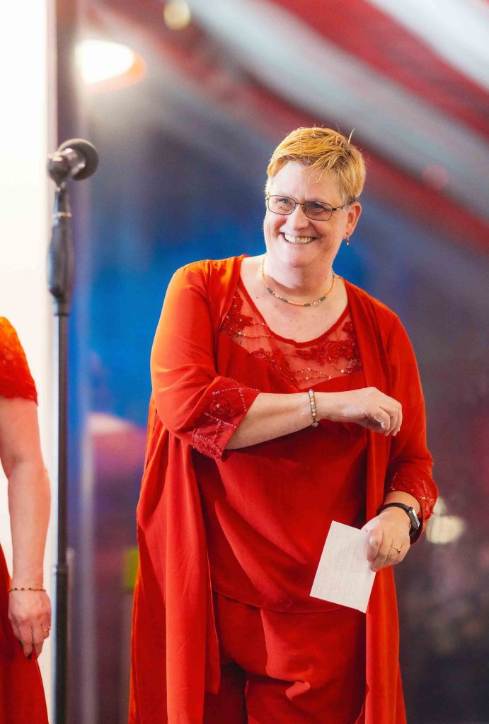 A person wearing glasses and a red outfit smiles while standing near a microphone and holding a piece of paper at a wedding reception in Baltimore.