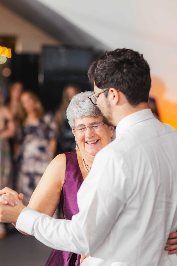 An older woman in a purple dress and a younger man in a white shirt are smiling and dancing together at a wedding reception.