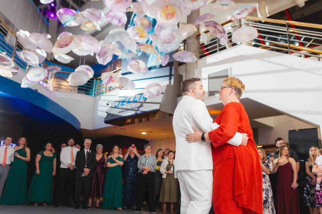 Two people dance together in the center of a room during a wedding reception while a group of guests watch; hanging decorations and a staircase are visible in the background.