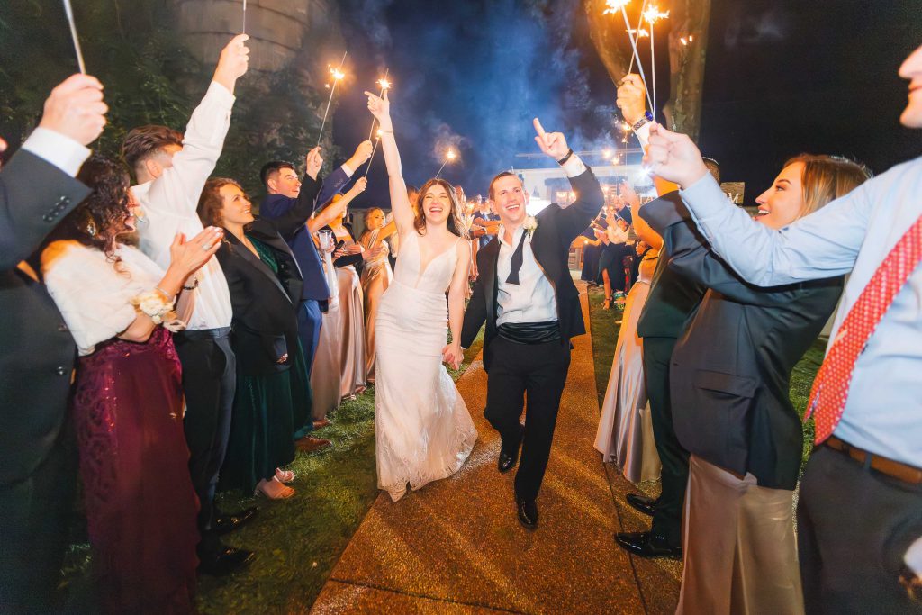 A bride and groom walk hand-in-hand down a path at The Barns at Hamilton Station, smiling and celebrating, surrounded by guests holding sparklers at night.