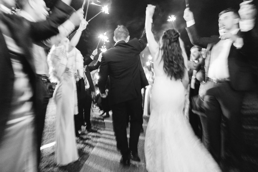 A black-and-white photo of a bride and groom walking through a crowd holding sparklers, captured from behind at their wedding reception. Guests are celebrating, some with arms raised and sparklers lit, creating a magical atmosphere at The Barns at Hamilton Station.