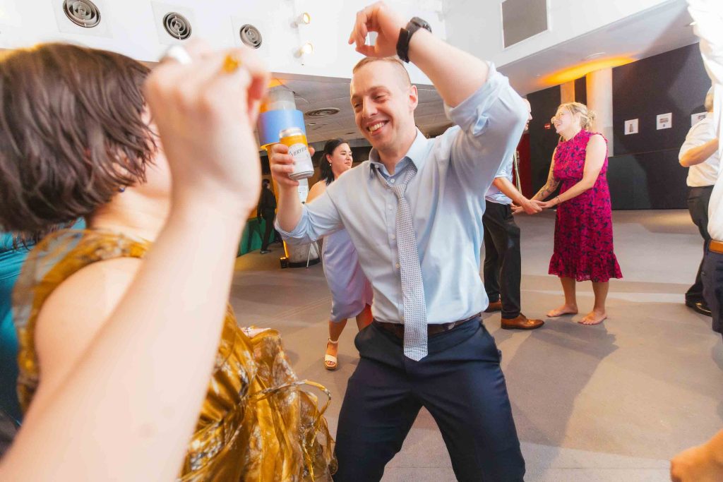 A man in a light blue shirt dances with a drink in hand at an indoor Baltimore wedding, surrounded by other guests who are also socializing and dancing.