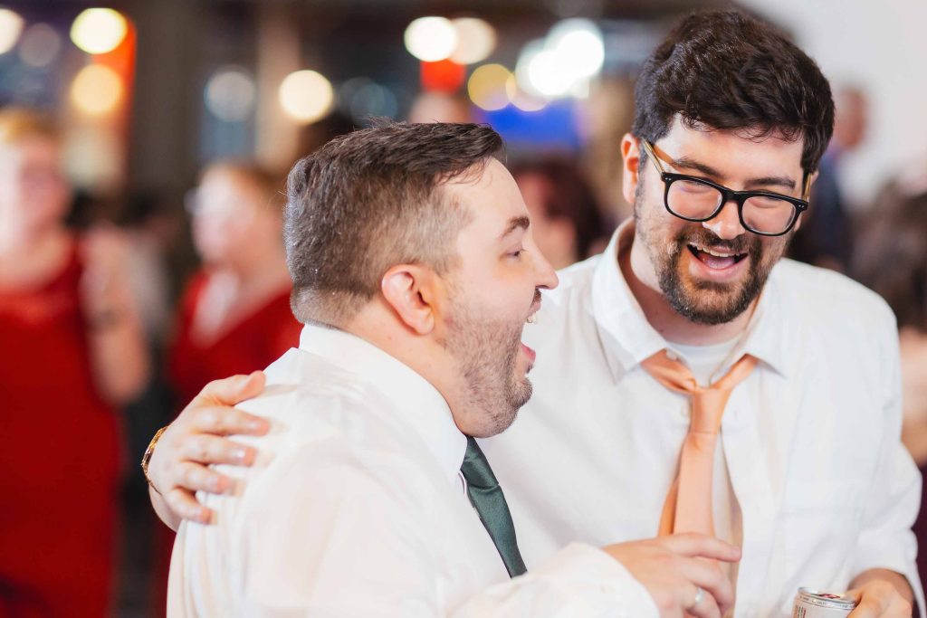 Two men in dress shirts and ties share a laugh at a reception, one with his arm around the other, with a blurred crowd in the background at the National Aquarium in Baltimore.