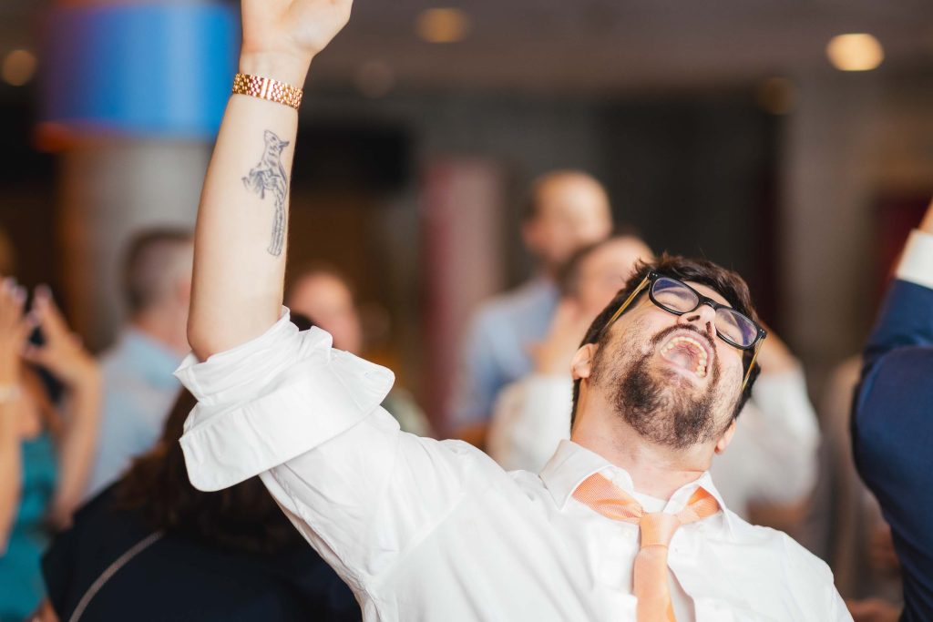 A man wearing glasses and a white shirt with a peach tie raises his arm and tilts his head back, appearing to celebrate at a lively reception in Baltimore’s National Aquarium.
