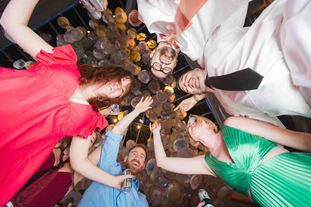 Five people in colorful outfits stand in a circle at a wedding reception, smiling and raising their hands while looking down at the camera; hanging lights are visible above them.
