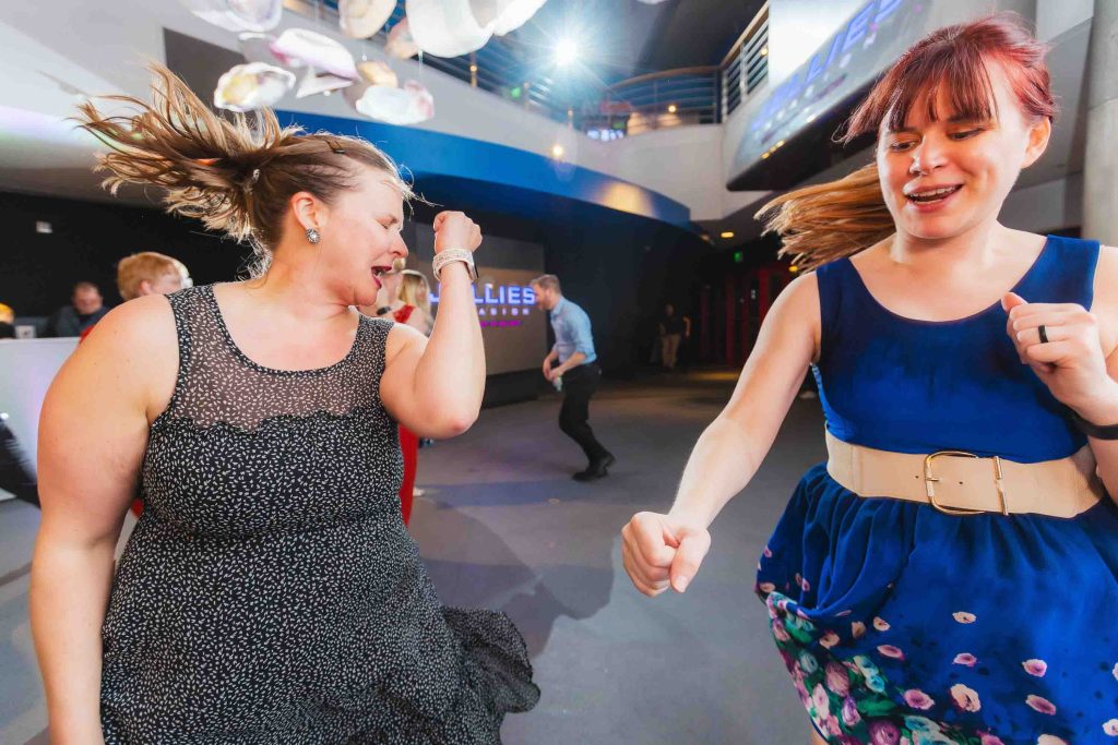 Two women are dancing enthusiastically indoors at a lively reception, swinging their arms and smiling. Other people are visible in the background under bright lighting, capturing the vibrant spirit of Baltimore.