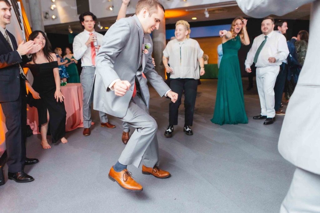 A man in a gray suit dances enthusiastically at a wedding reception while people around him watch, smile, and clap.