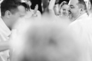 A group of people dancing and celebrating at a wedding reception indoors in Baltimore; the image is in black and white with a blurred foreground and two men visible in focus.