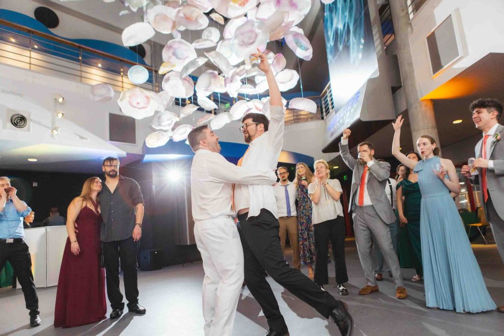 Two men celebrate and embrace on a dance floor surrounded by clapping and cheering guests in formal attire, under hanging decorative elements at a modern Baltimore wedding venue.