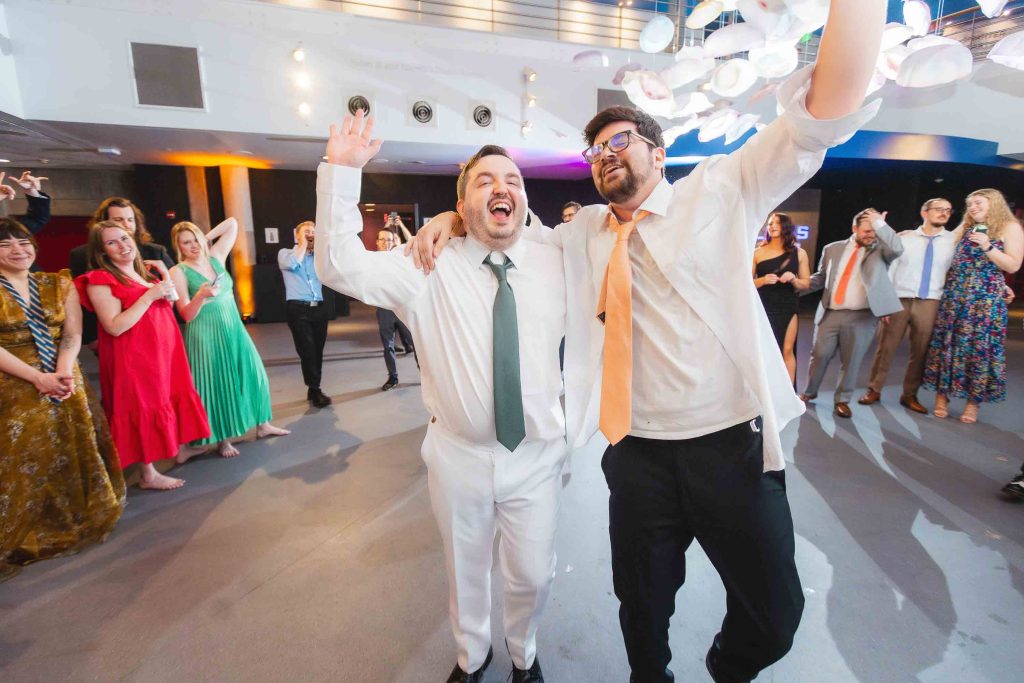 Two men with raised arms smile and celebrate on the dance floor at a Baltimore wedding reception, surrounded by applauding guests in formal attire at an indoor event.
