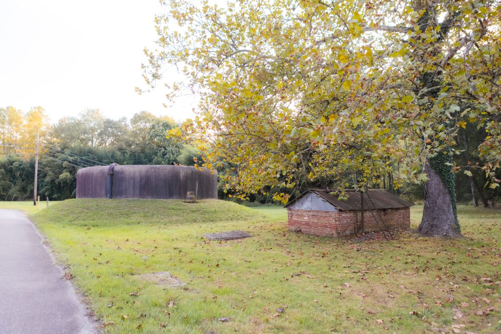 Grassy area with a large, round concrete structure reminiscent of Annapolis Waterworks Park and a small, brick shed surrounded by trees. A road runs alongside.