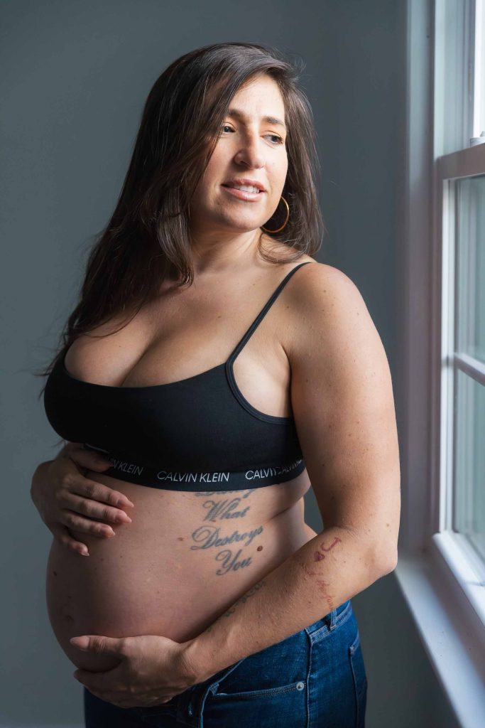 A maternity portrait of a pregnant woman in a black Calvin Klein bra and jeans stands by a window, holding her belly and looking outside in a studio style setting.