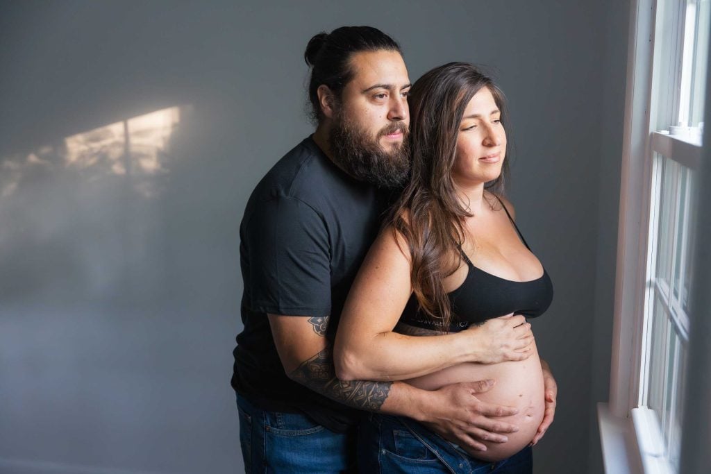 A man stands behind a pregnant woman, both with hands on her belly, looking out a window in a softly lit, studio style maternity room.