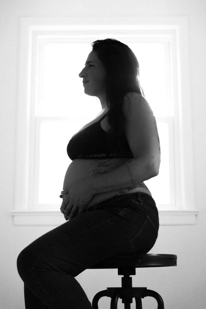 A studio style maternity portrait of a pregnant woman sitting sideways on a stool, gently holding her belly, with a bright window in the background.