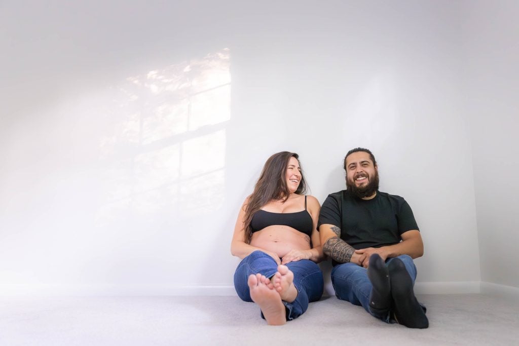 A pregnant woman and a man sit barefoot on the floor, leaning against a white wall, smiling at each other in this cozy indoor studio style portrait.