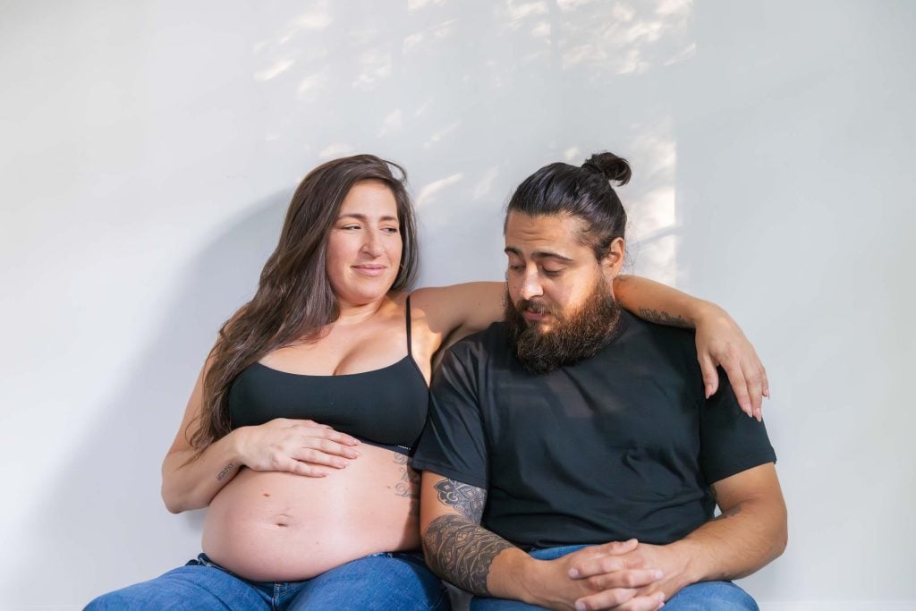 A pregnant woman sits with her arm around a man, both wearing black tops and jeans, in a Studio Style maternity portrait against a plain white background.