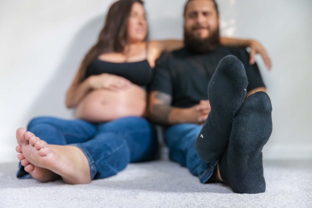 A maternity portrait features a pregnant woman and a man sitting on the floor, legs extended forward; their faces are out of focus while their feet take prominence in this studio style composition.