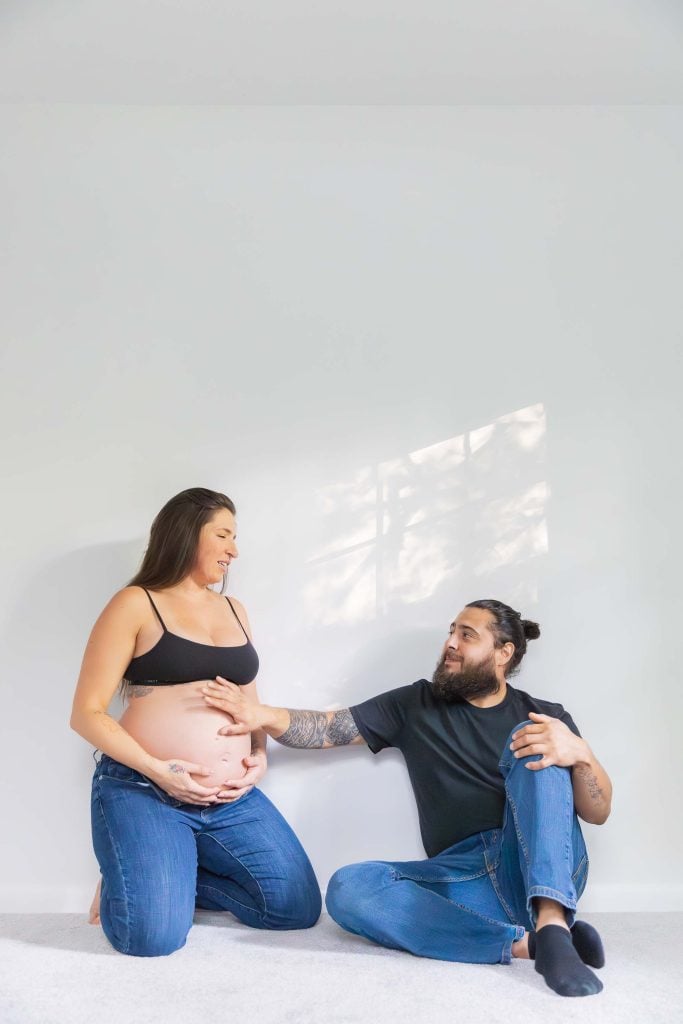 An indoor maternity portrait shows a pregnant woman sitting on the floor next to a man, both in jeans and black tops. The man gently rests his hand on her belly as they pose against a plain white wall.