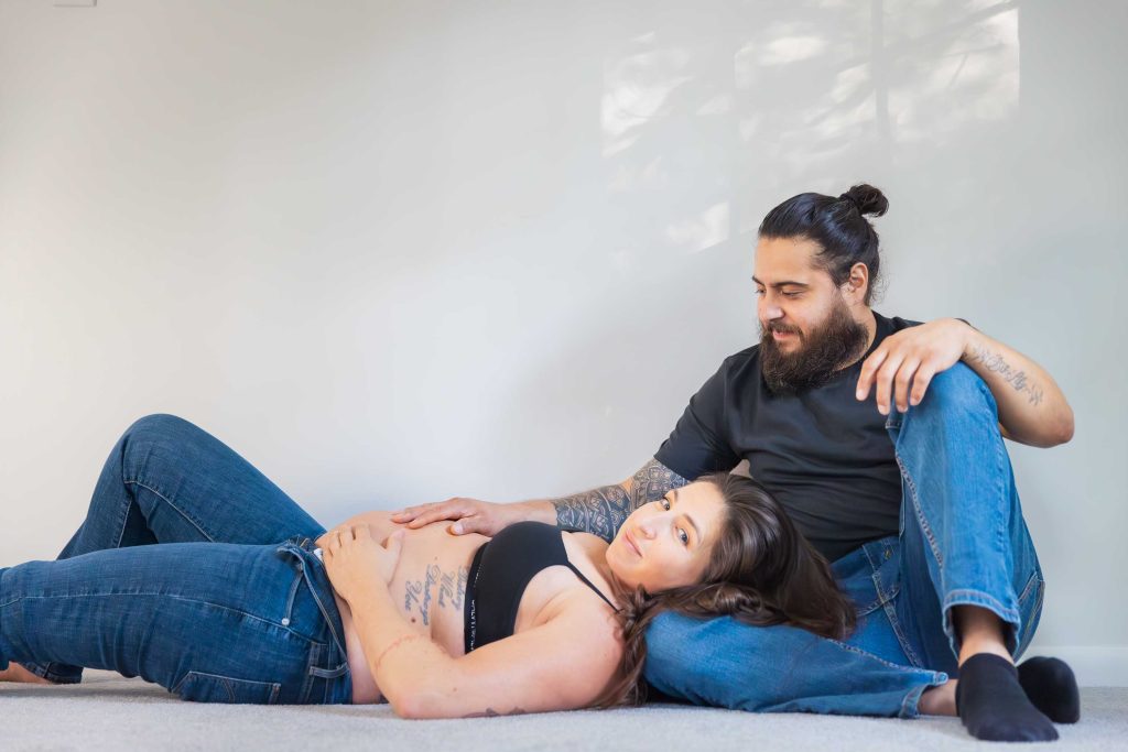 An intimate indoor maternity portrait shows a pregnant woman lying on the floor with her head on a man's lap, both dressed in jeans and casual tops, set against a plain light-colored wall.