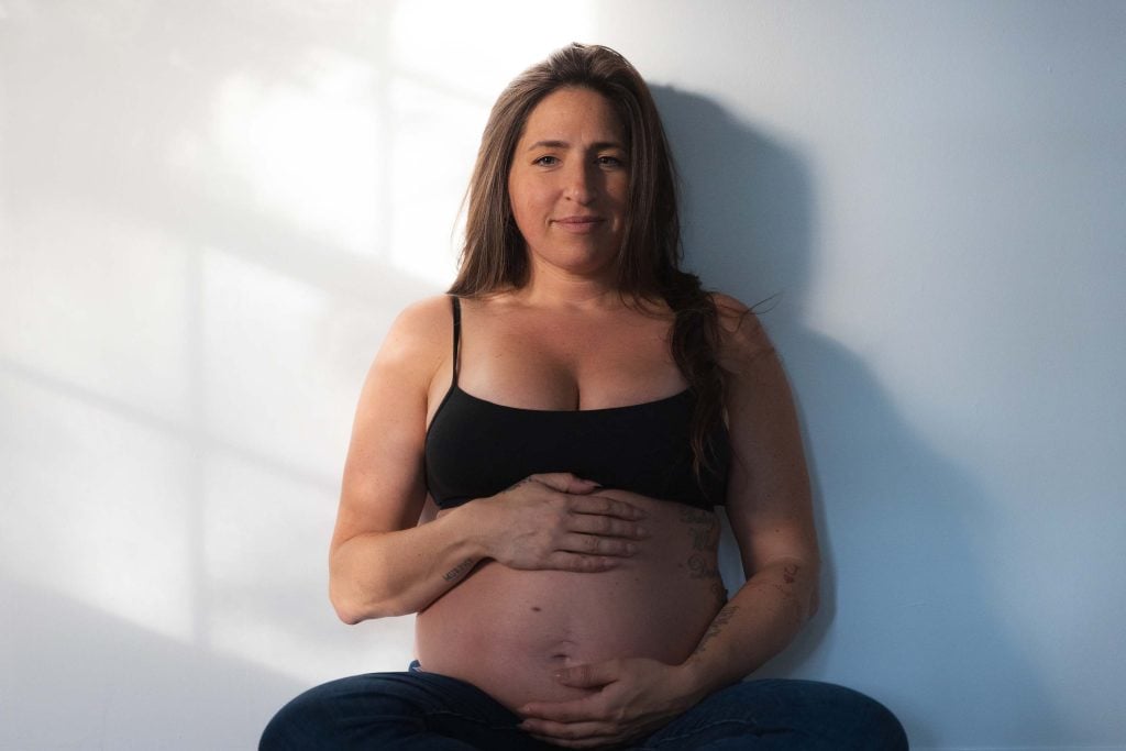 This indoor maternity portrait shows a pregnant woman in a black sports bra sitting against a white wall, resting her hands on her bare belly as sunlight casts soft shadows around her.