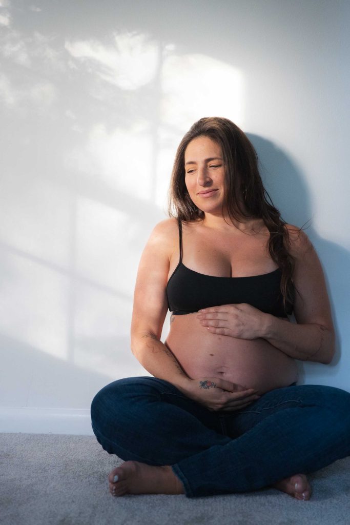 An indoor maternity portrait of a pregnant woman in a black bra and jeans, sitting cross-legged on the floor with her hands on her bare belly as sunlight casts shadows on the wall behind her.