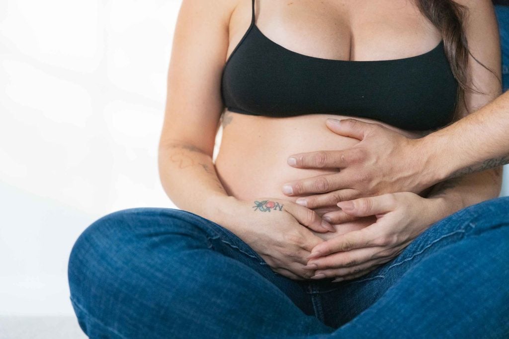 A pregnant person in a black sports bra and jeans sits cross-legged indoors, with another person's hands gently resting on their bare belly—a tender maternity portrait.