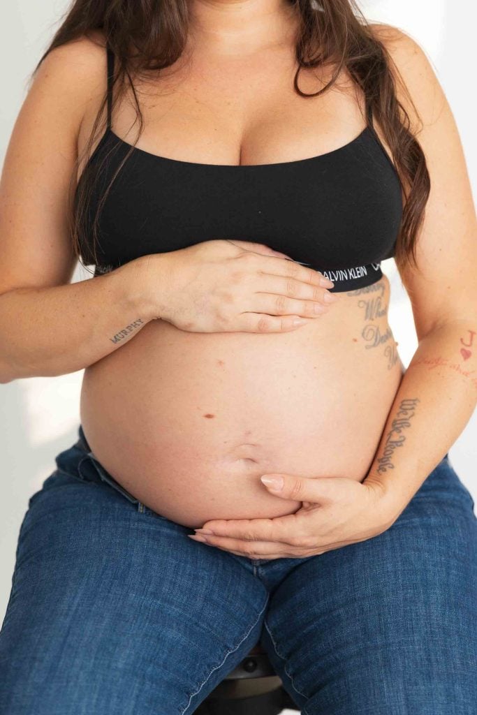 A pregnant person wearing a black bra and blue jeans sits indoors with hands resting on their bare belly, capturing a serene maternity moment in studio style.