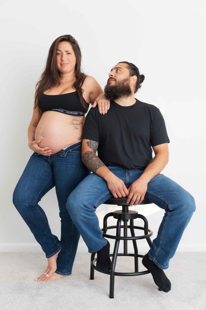 A pregnant woman stands with her hand on her belly next to a seated, bearded man with tattoos. Both are dressed casually in jeans and black tops, captured in a classic indoor studio style against a plain white background.