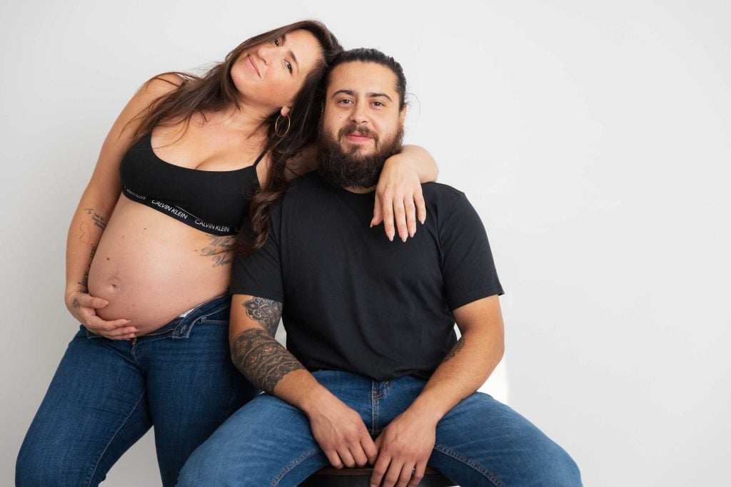 A pregnant woman in a black top and jeans stands beside a seated man in a black t-shirt and jeans, both posing for maternity portraits in a classic studio style against a plain white background.