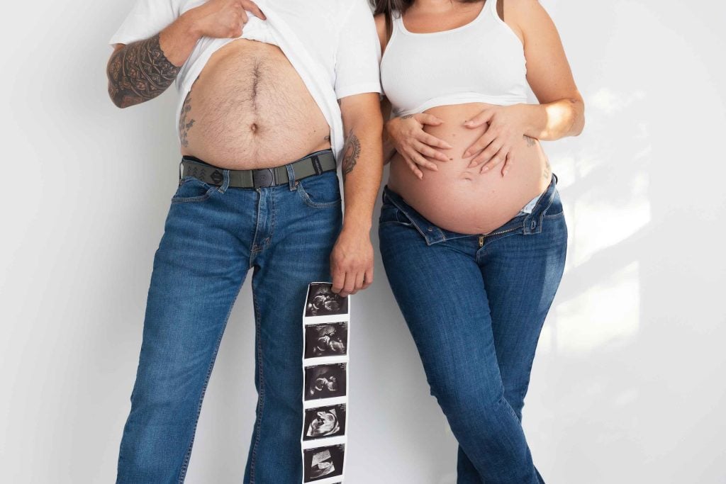 A studio style maternity portrait featuring a pregnant woman and a man in jeans and white tops; the man lifts his shirt as they stand side by side, holding a strip of ultrasound images.
