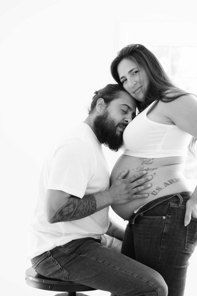 A man with tattoos rests his head and hands on the belly of a pregnant woman in this intimate indoor maternity portrait, both dressed in casual white tops and jeans in a bright room.