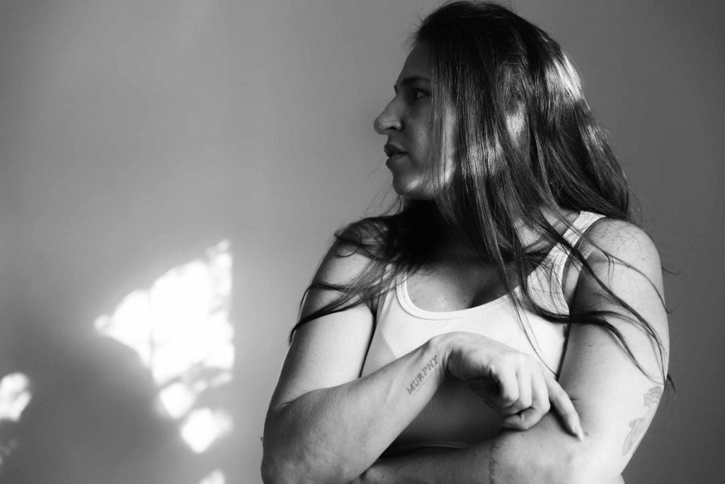 A woman with long hair, wearing a sleeveless shirt, stands with arms crossed and looks to the right in an indoor room lit by natural light, creating a serene portrait.