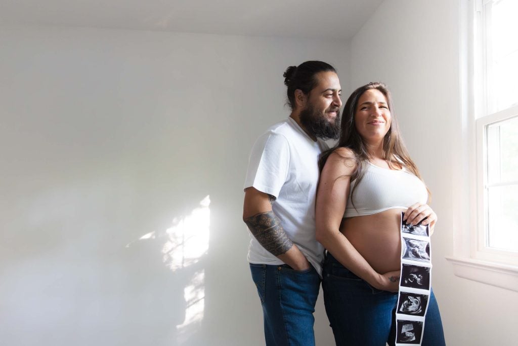A pregnant woman holds ultrasound photos while standing by a window with a man beside her; both are casually dressed and smiling in this warm indoor maternity portrait.
