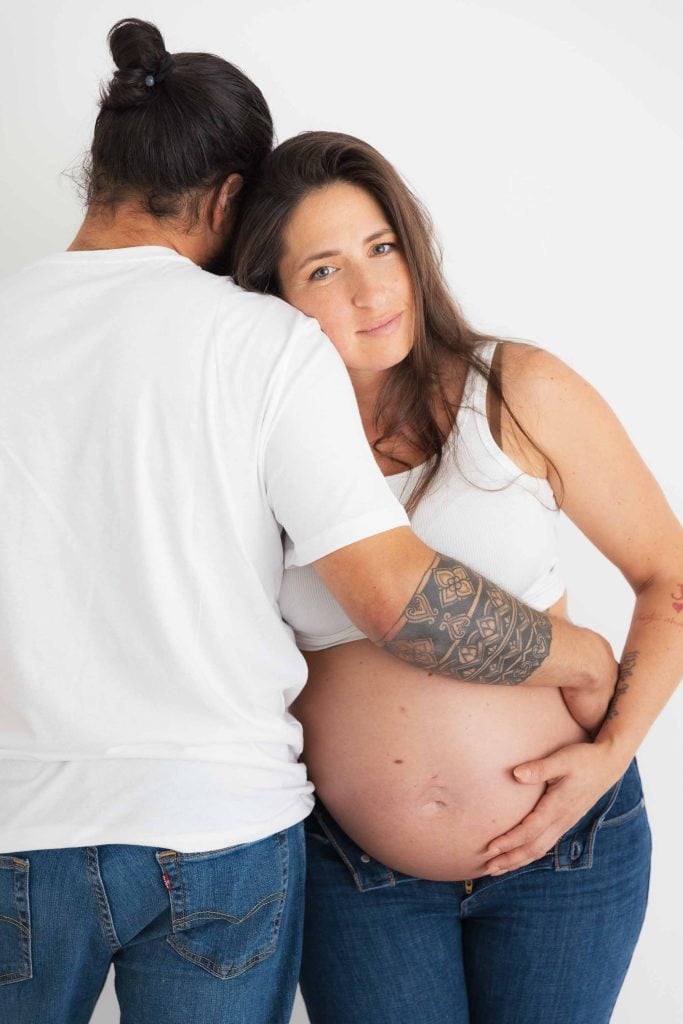 A pregnant woman in a white tank top and jeans stands next to a man in a white t-shirt and jeans, who embraces her with his tattooed arm in a cozy indoor, studio-style maternity scene.