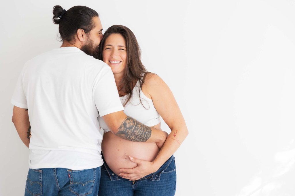 A man embraces and kisses a smiling pregnant woman on the cheek in this intimate indoor portrait. Both wear white shirts and jeans, and she warmly cradles her bare belly.