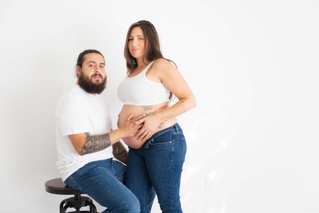 A man sits on a stool next to a pregnant woman standing, both in matching white tops and jeans. His hand rests on her bare belly as they face the camera for classic studio style portraits against a plain white background.