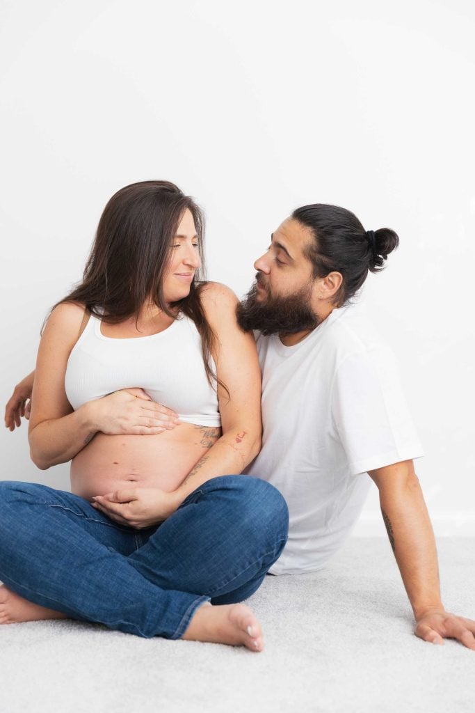 A pregnant woman sits on the floor next to a man in a studio-style indoor setting. She rests a hand on her belly, and both wear casual white tops and jeans against a plain white background.