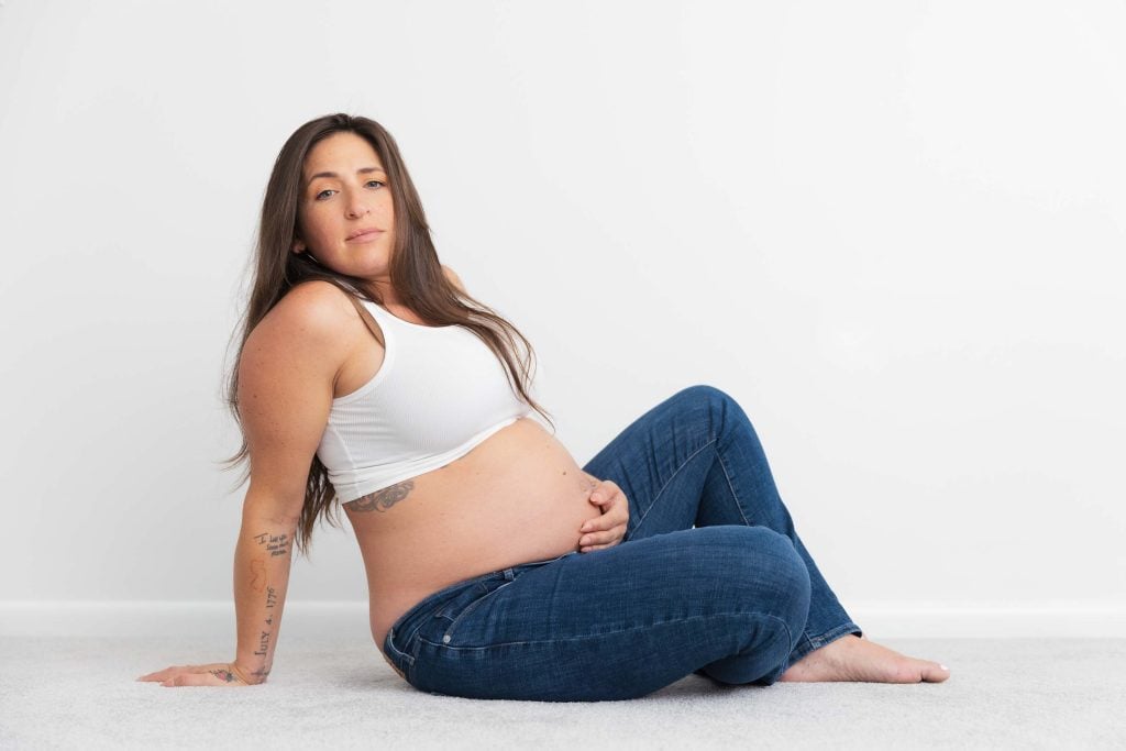A maternity studio style portrait of a pregnant woman in a white tank top and blue jeans, sitting on the floor against a white background, looking at the camera and resting her hand on her belly.