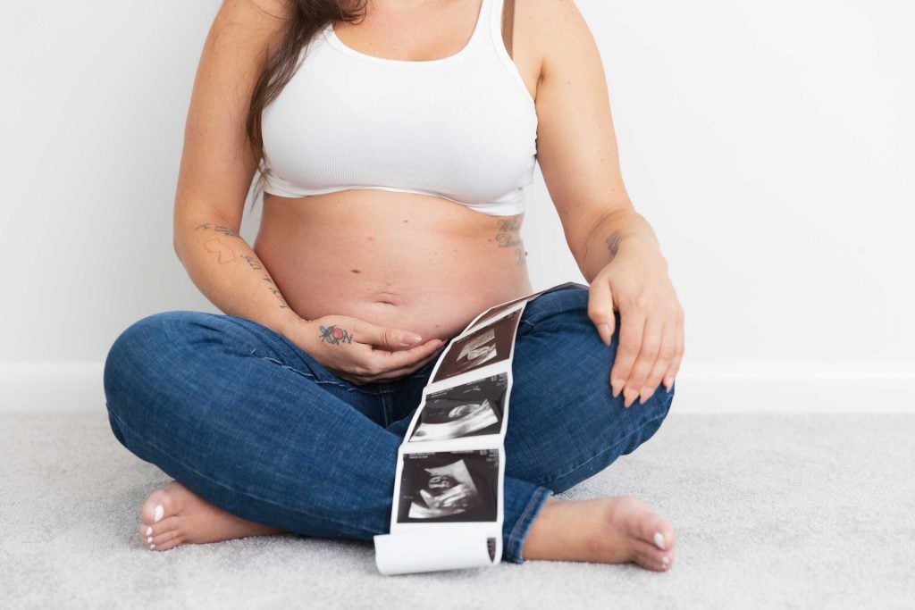 Maternity portraits: A pregnant person in a white top and jeans sits cross-legged indoors, holding their belly and a strip of ultrasound images.