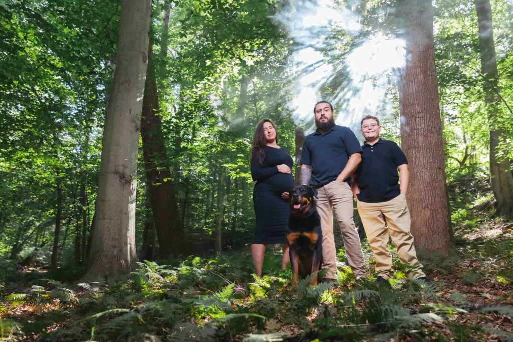 Three people and a dog stand together in a sunlit forest, with rays of light streaming through the trees, creating a beautiful outdoor portrait.